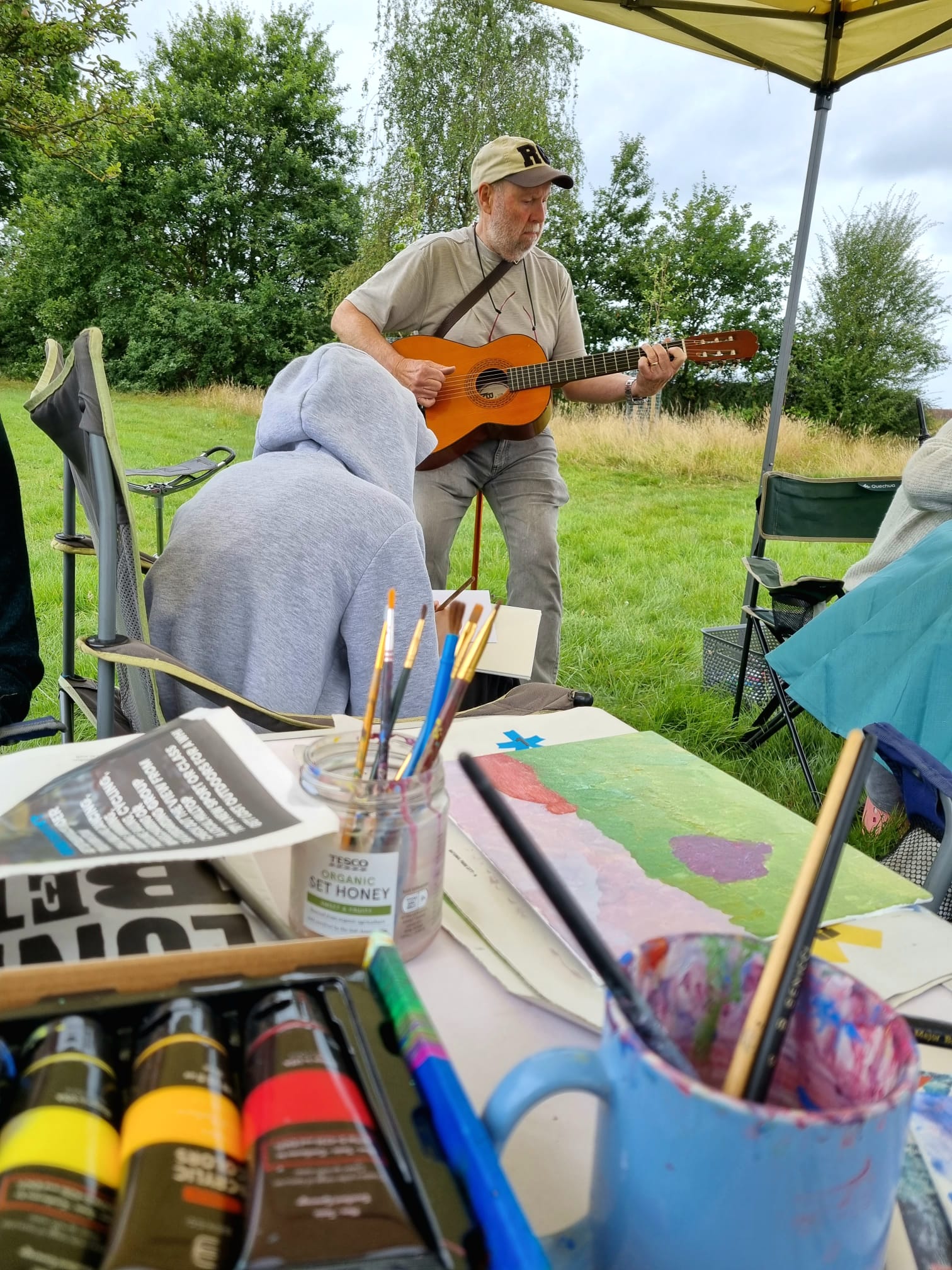 A man with a beard sits on a stick stool playing his guitar. There are bushes and grass in the background. In the foreground are paints and brushes and a girl painting.