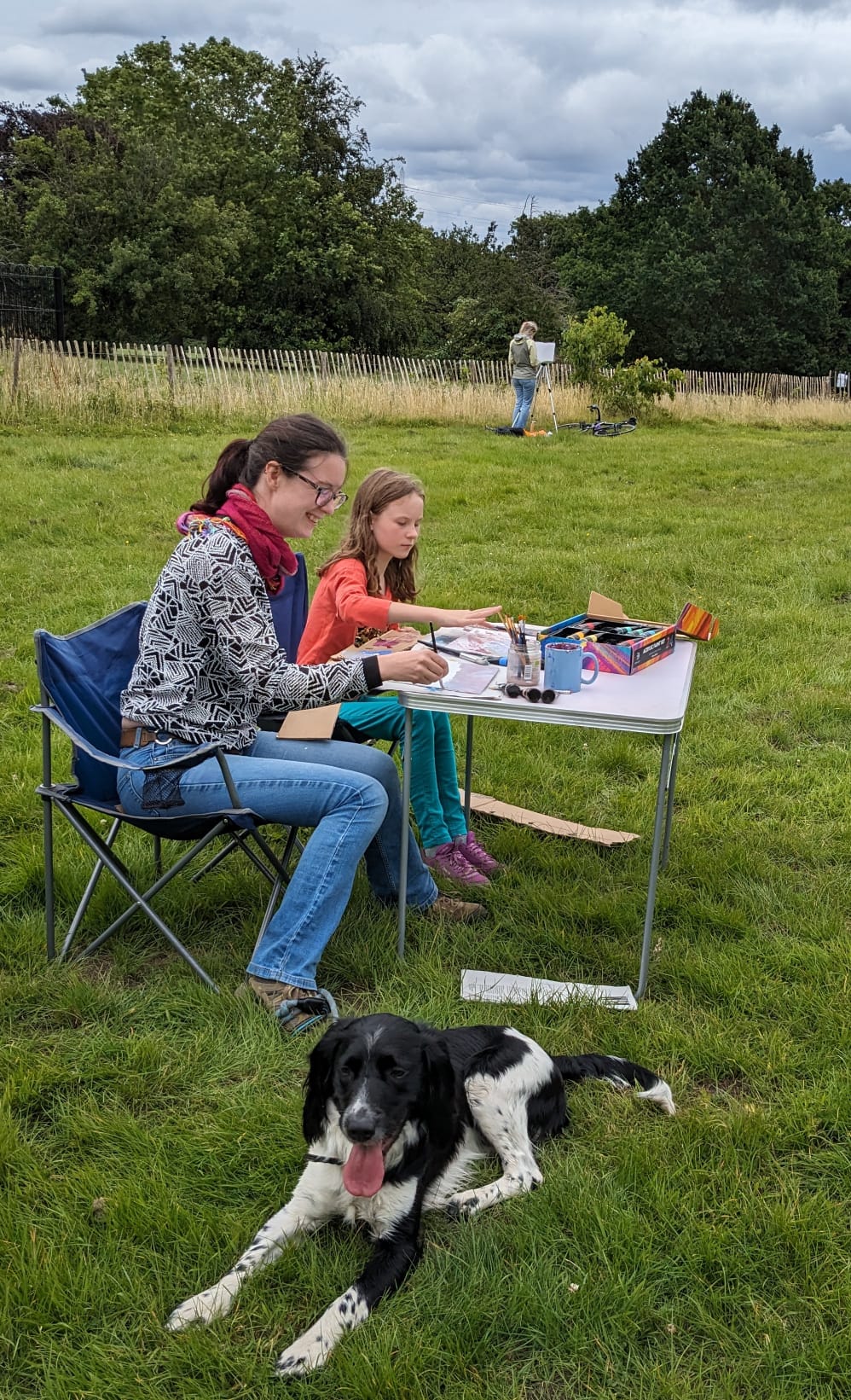 A mother and daughter paint sitting at a table while a black and white dog lies on the grass facing the camera