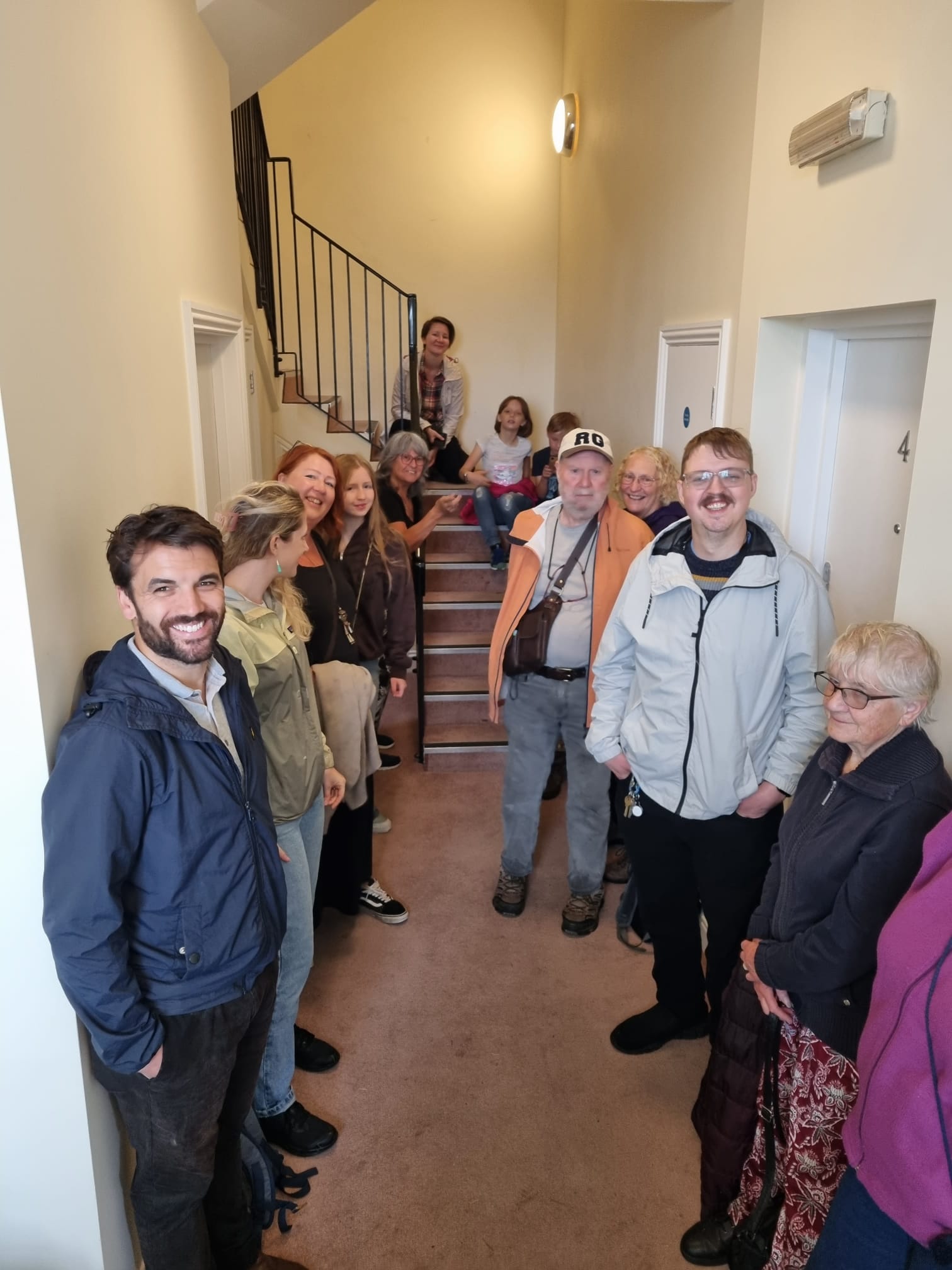 A group of 13 women and children smile to camera, standing by the walls in a hallway with stairs at the end. The children sit on the stairs with a grown up.