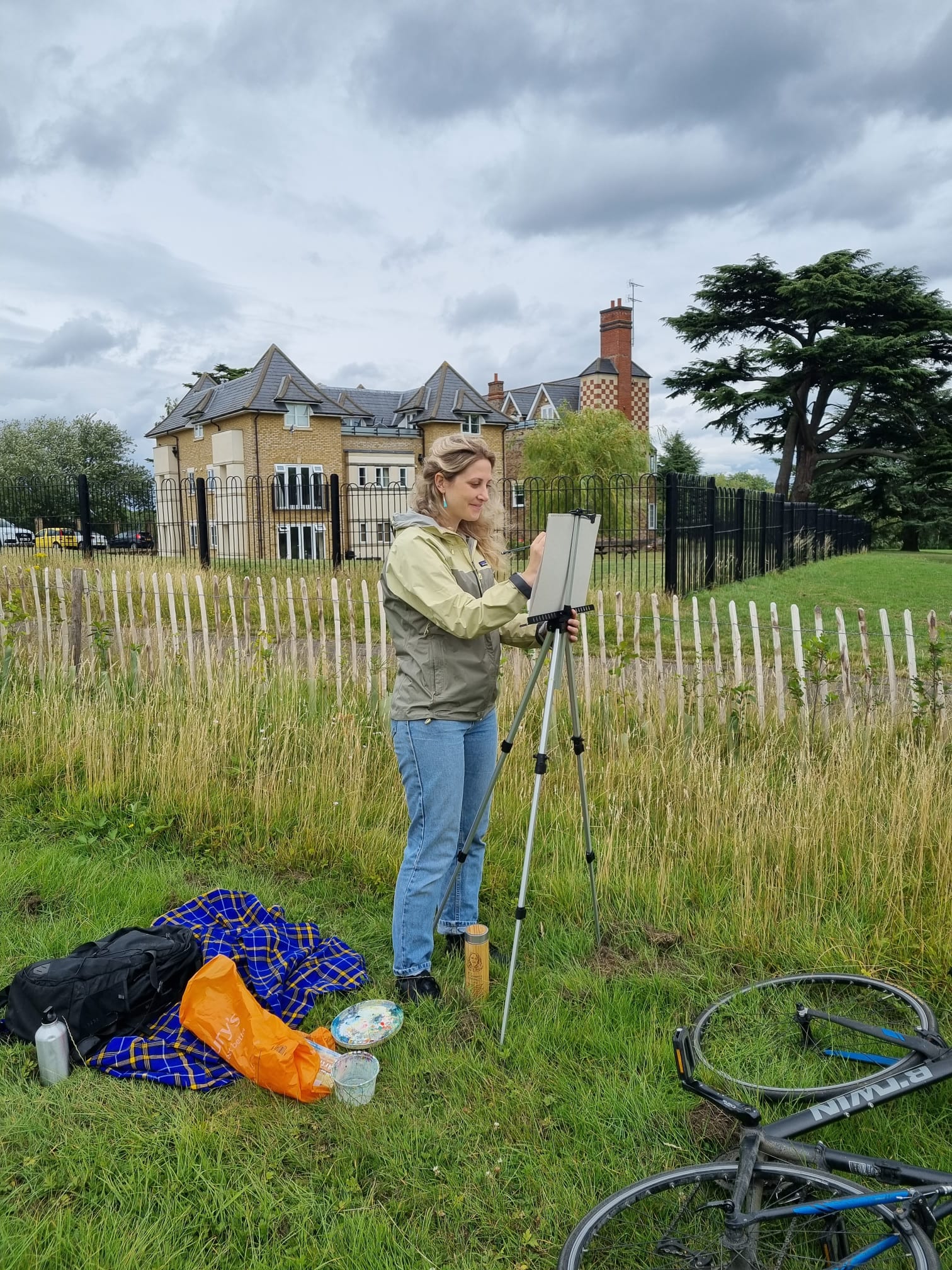 Gemma is blond and wears jeans and a yellow waterproof stands facing her easel with her back to Bishopsford House, 2 blocks of flats made of brick in the middle of the park.