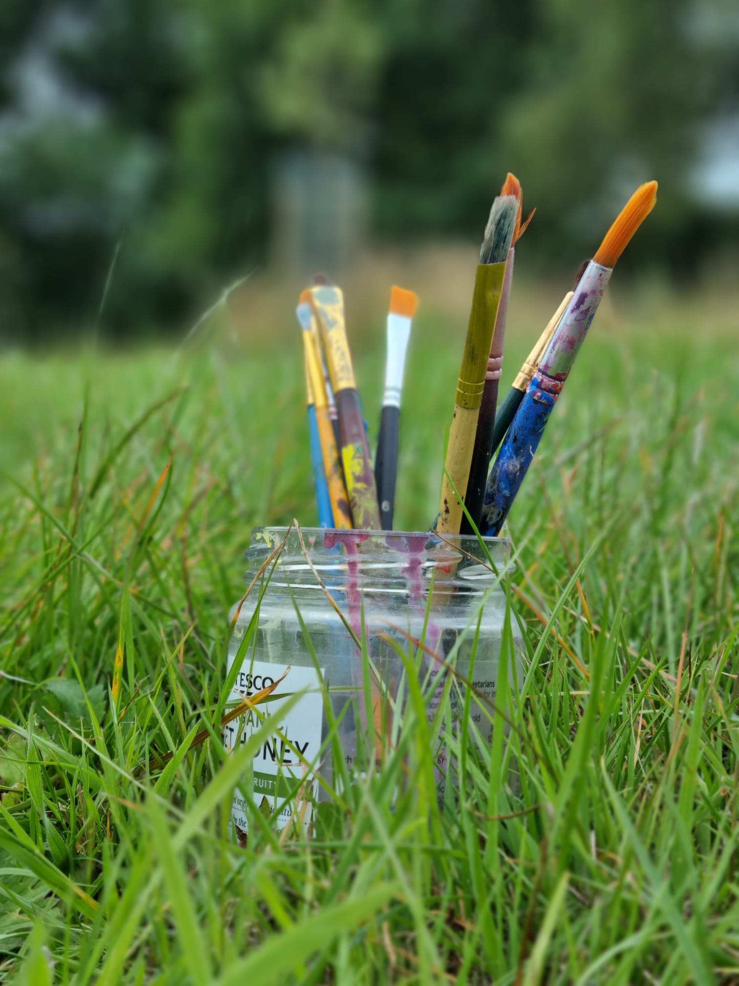 A glass jar sits on the grass with 7 different paintbrushes in it