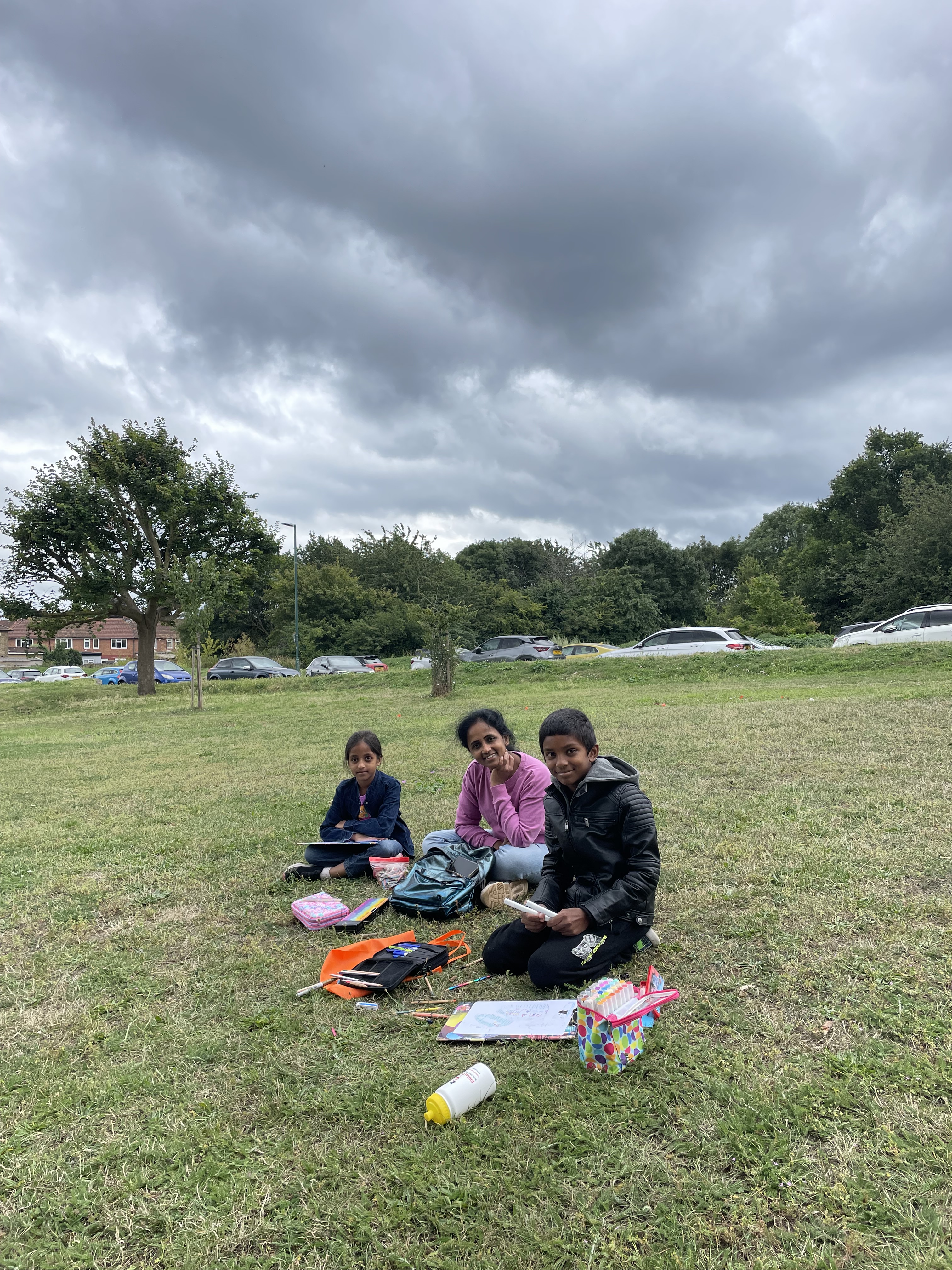 A young family sit on the grass with art materials under a grey sky