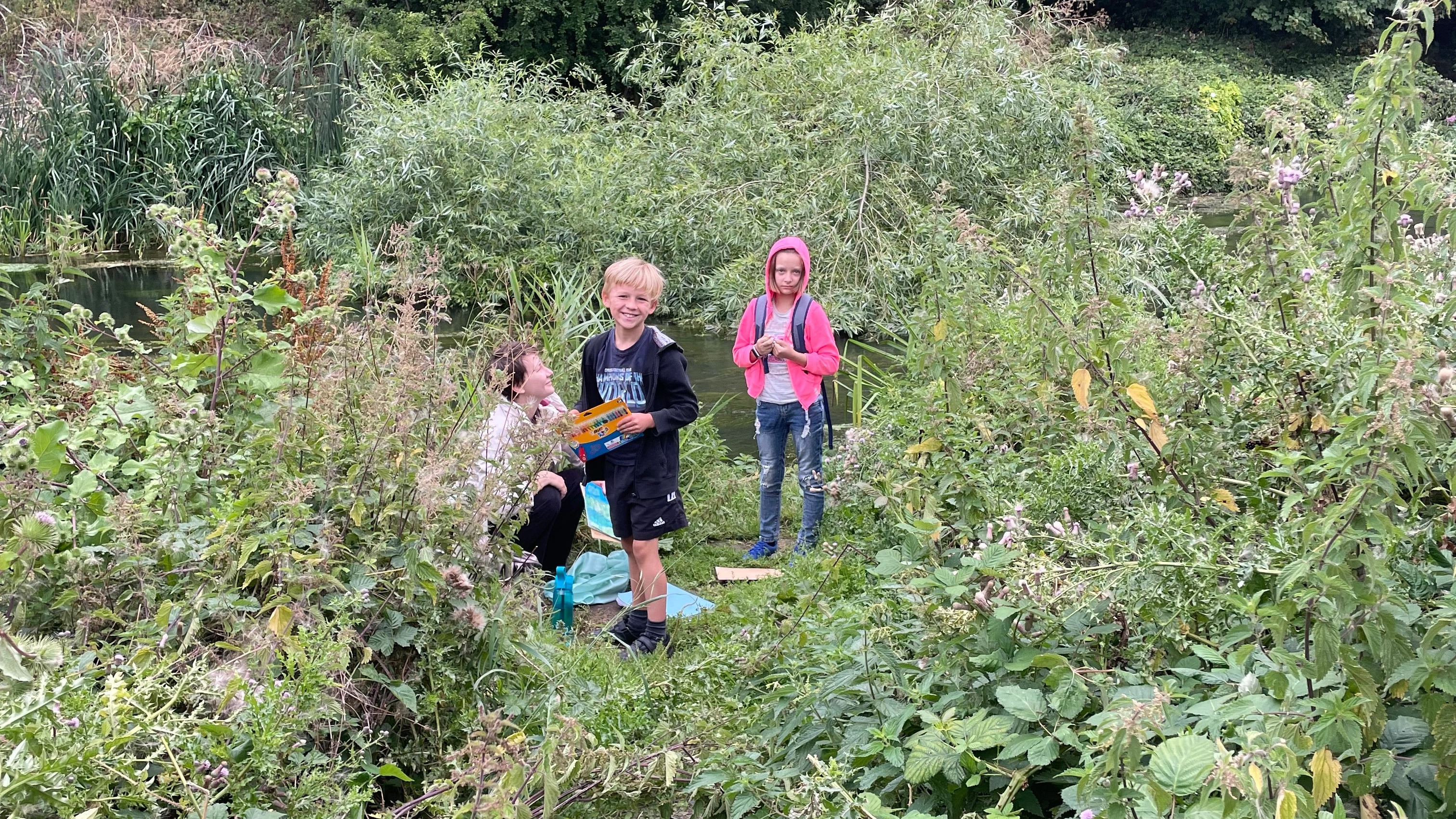 A woman sits with a little boy and girl standing with crayons among the green vegetation on the bank of the river Wandle