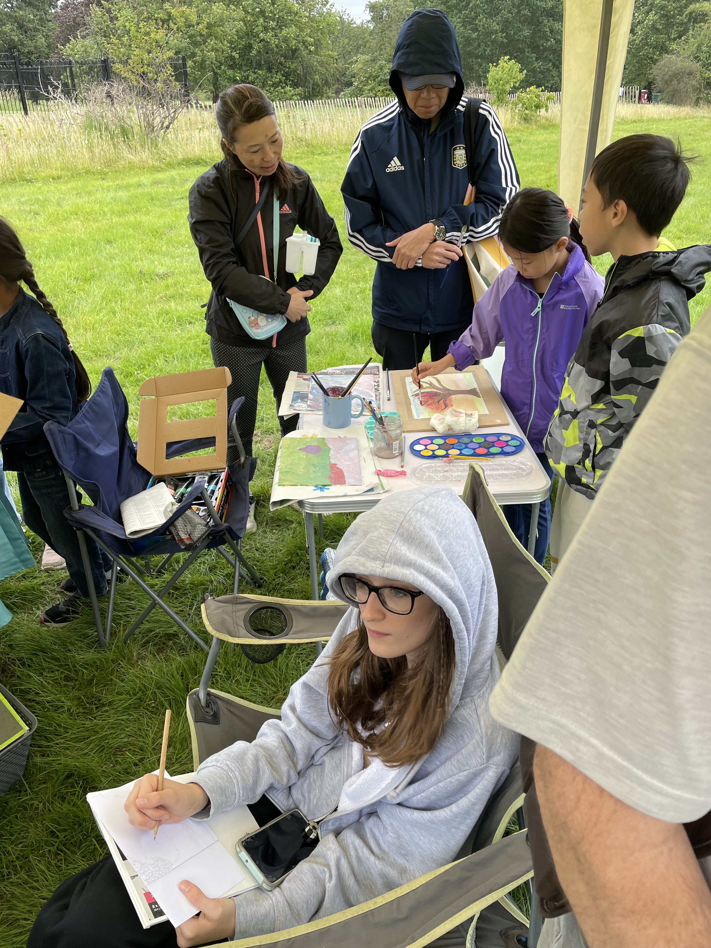 A family stand with their two children painting at the back of a marquee. In the foreground a young girl draws.