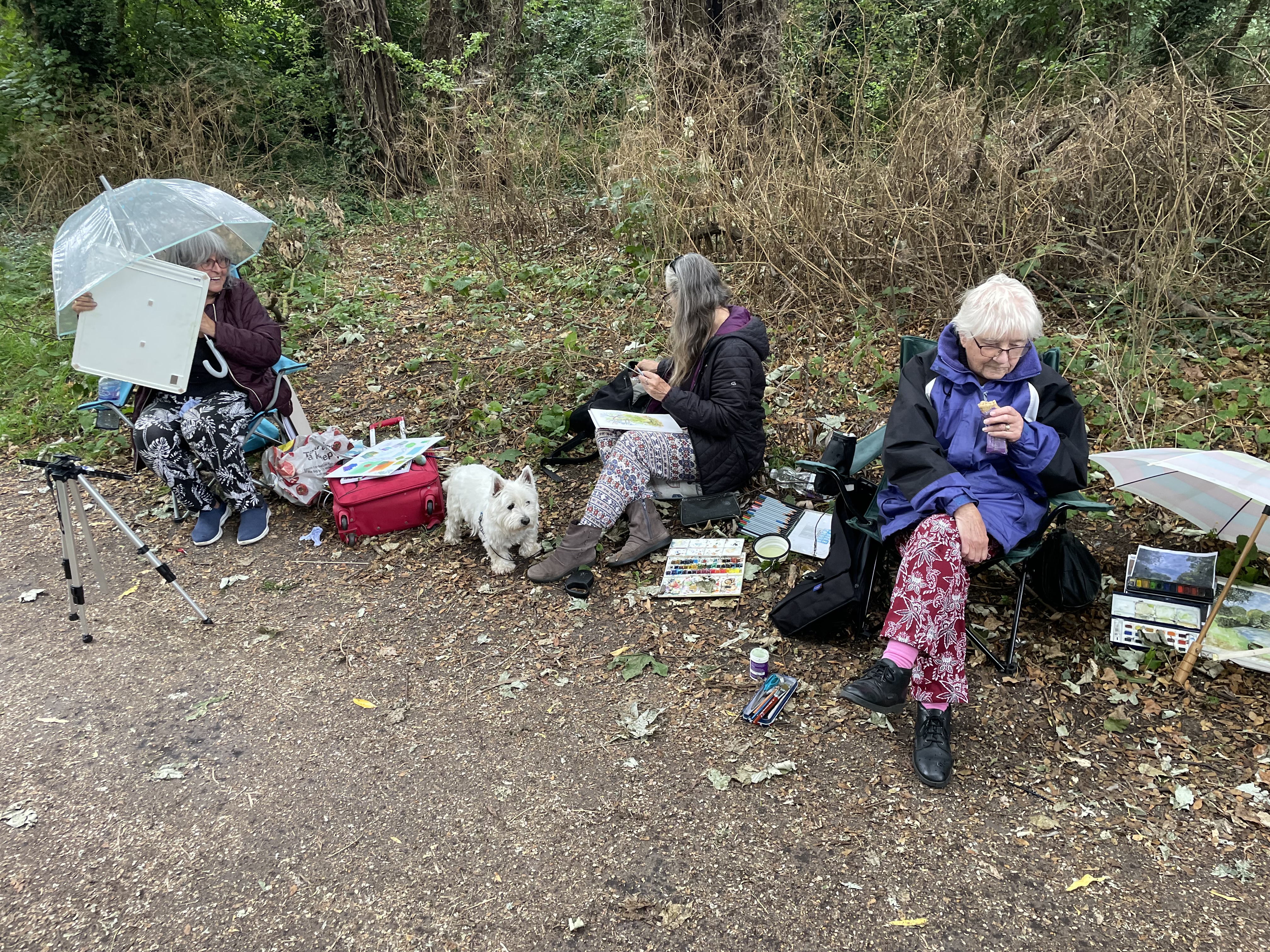 Three artists sit in a line along the river path with a small white scottie dog