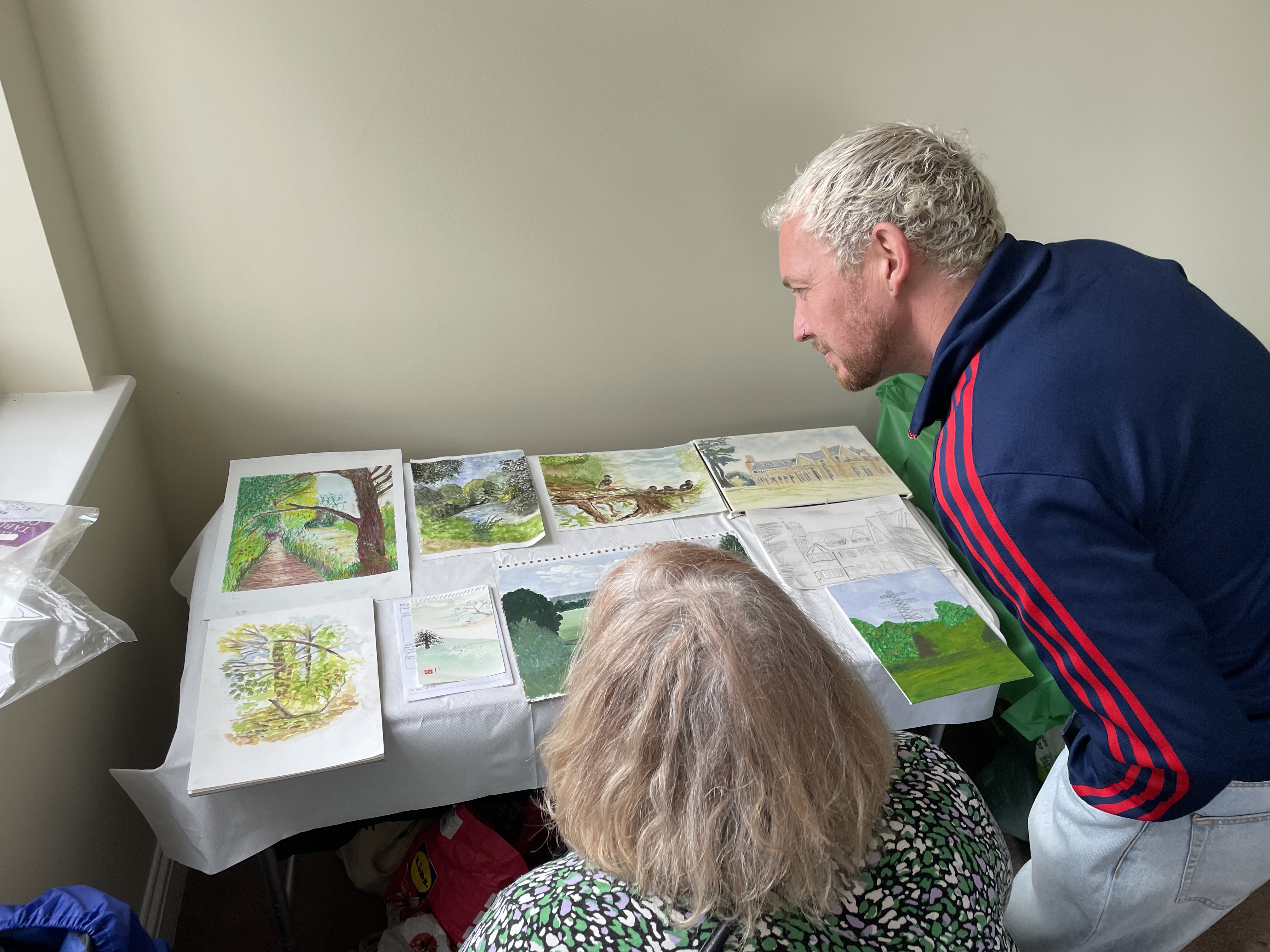 Judges Rhys and Linda look at 9 paintings and drawings laid out on a table. Rhys bends over he is wearing a blue jacked with orange piping on the sleeves. Linda is sitting down in her wheelchair you can see the top of her head and her black and green dress.