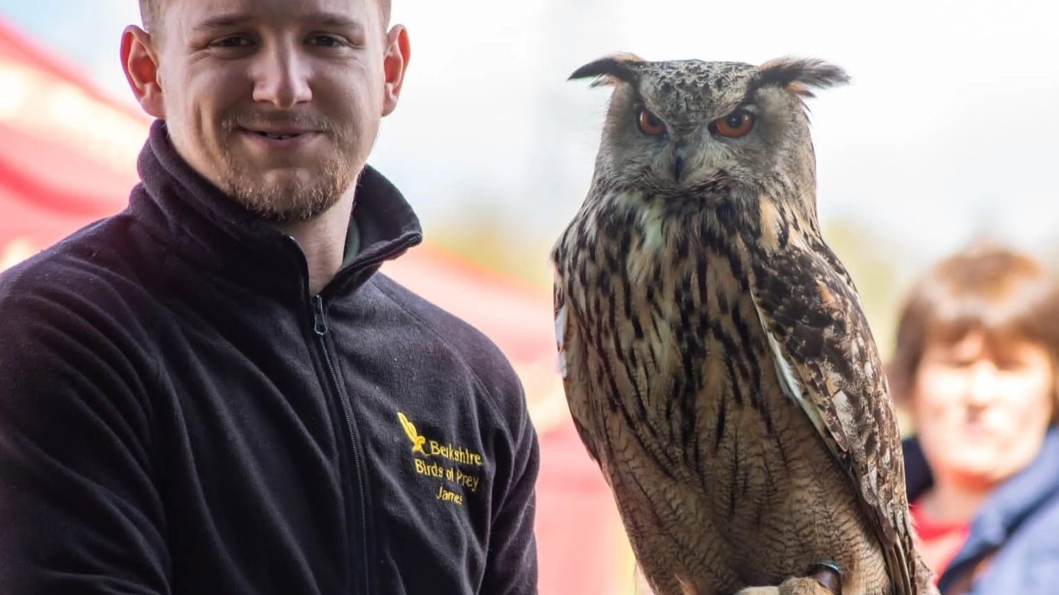 A smiling ginger man in a black fleece holds a brown specked european owl. It is about 4 foot tall and has orange eyes and feathery tufts on either side of its head.