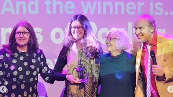 Lady with spotty dress presents volunteers Isobel and Sandra with comedian Tim Vine on the right. Above them lettering reads And the winners are...