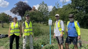 A blue signpost is planted in a flower bed. Four people in high vis stand around it