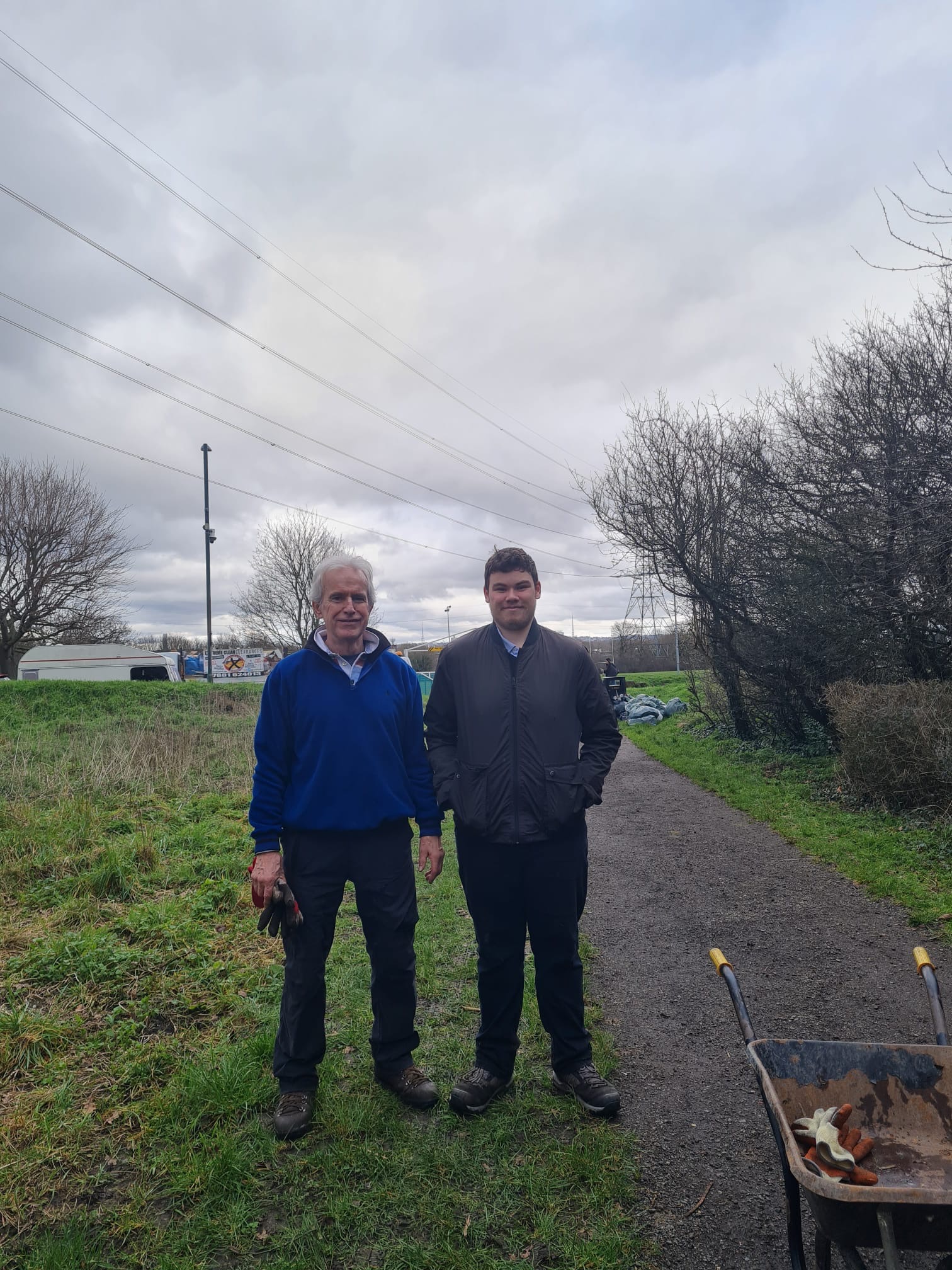 Steve and Steve stand on the path in Revesby Wood against a grey sky One steve has white hair and a blue jacket the other has black hair and a black jacket