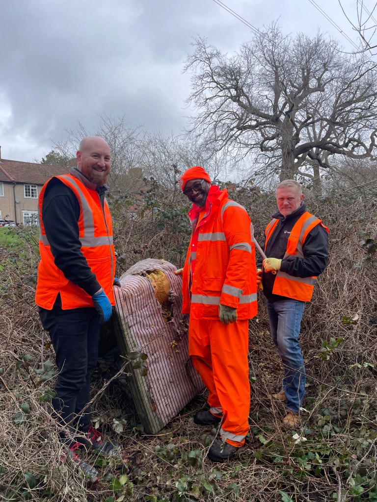 3 Veolia volunteers in orange high vis drag a mattress out of a thick copse of brambles
