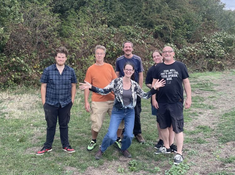 Steve, Spike, Nick, Matt and Charlie stand in front of a woodland behind Isobel who is crouching and smiling with arms spread wide. Spike is pulling a face.