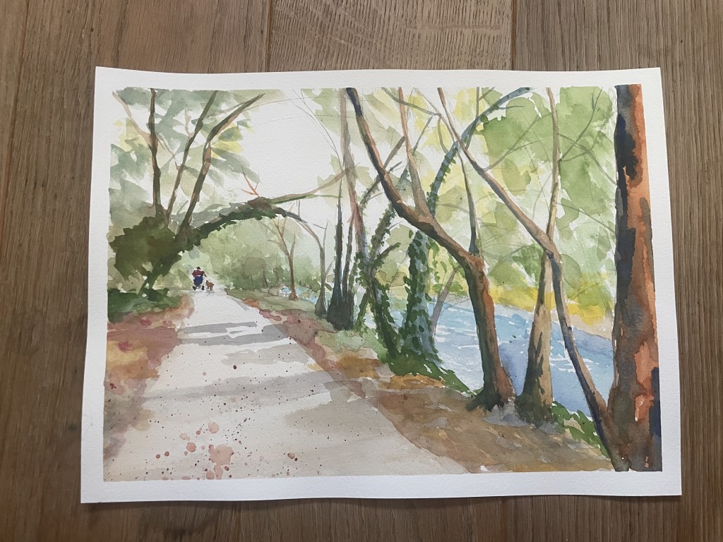 Elegant ivy covered trees frame a path by the river. In the distance is a the back of a person in a mobility scooter taking their dog for a walk.