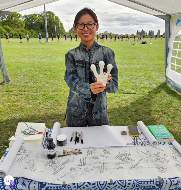 Ava stands behind a table with long strips of paper and ink and pens. She holds a plaster hand with 3 toes and is smiling.